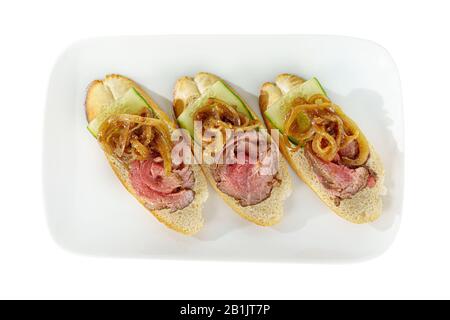 Sandwichs froids en hors-d'œuvre avec rôti de bœuf mariné, oignons marinés et concombre sur une plaque carrée, vue sur le fond blanc isolé d'en haut pour le Banque D'Images