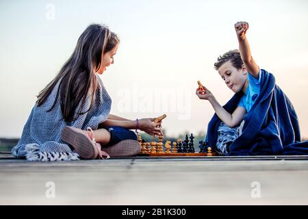 Deux jeunes petits amis mignons, un garçon et une fille qui s'amusent en jouant aux échecs assis près du lac le soir. Les enfants jouent.geste gagnant. Ami Banque D'Images