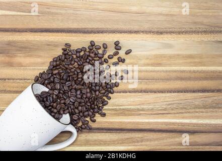 Vue de dessus d'un mug à café blanc Sur Son Côté, En Bas à gauche du cadre, avec des grains de café organiques Débordant sur une table En bois Rustique Banque D'Images