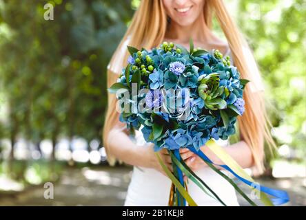 Crop souriant jeune fille blonde à poil long tenant un beau bouquet élégant de fleurs vertes et bleues avec des rubans tout en se tenant dans le parc avec une purée Banque D'Images
