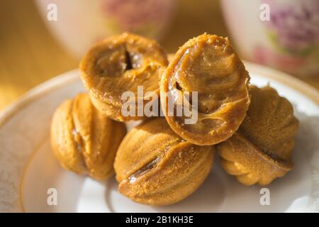 Biscuits en noyer avec lait condensé dans la soucoupe blanche. Un ouvert dans la moitié. Bonbons sur la table ensoleillée. Tasses à thé en arrière-plan flou. Banque D'Images