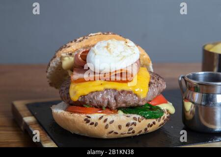 Hamburger de bœuf maison, oeuf poché, sauce hollandaise et bacon croustillant et fromage cheddar sur planche en bois. Image verticale Banque D'Images