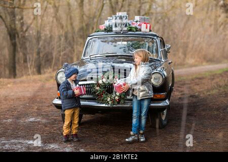 Charmante fille et garçon élégante donnant des cadeaux à l'autre debout près de la voiture noire rétro avec des boîtes sur le toit dans le parc d'hiver Banque D'Images