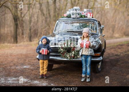 Charmante fille et garçon élégante donnant des cadeaux à l'autre debout près de la voiture noire rétro avec des boîtes sur le toit dans le parc d'hiver Banque D'Images