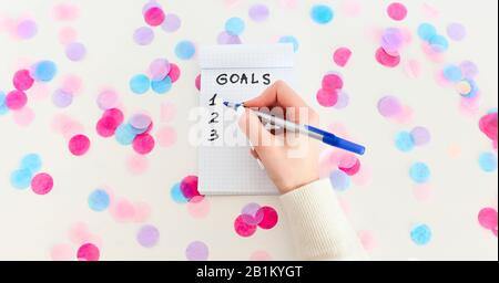 Main féminine avec stylo écriture des buts de nouvel an dans un bloc-notes vide sur fond blanc avec confetti Banque D'Images