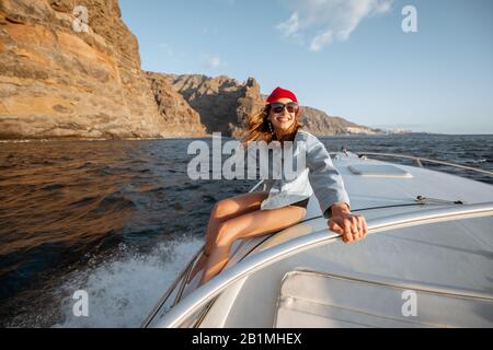 Jeune femme vêtue à la fois castiquement en veste et chapeau rouge en voyage maritime, naviguant sur un yacht dans l'océan orageux près d'une côte rocheuse. La poursuite sportive extrême de l'été Banque D'Images