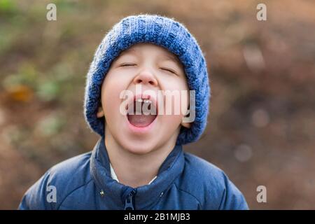 Petit garçon mignon dans une veste chaude bleue et bonnet tricoté souriant contre un parc flou par temps froid Banque D'Images