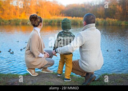 Père, mère et petit-fils par le lac chez les canards à l'automne Banque D'Images