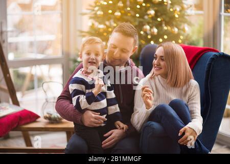 Joyeux parents et fils avec de délicieuses bonbons assis dans un fauteuil doux célébrant les vacances de Noël dans la salle de séjour avec un sapin brillant Banque D'Images