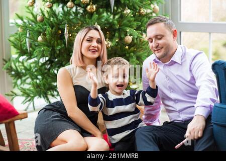 Joyeux parents et fils avec de délicieuses bonbons assis dans un fauteuil doux célébrant les vacances de Noël dans la salle de séjour avec un sapin brillant Banque D'Images