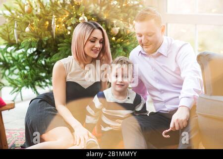 Joyeux parents et fils avec de délicieuses bonbons assis dans un fauteuil doux célébrant les vacances de Noël dans la salle de séjour avec un sapin brillant Banque D'Images