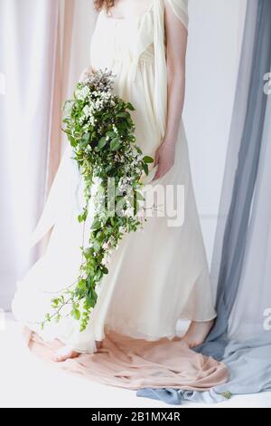 Chabillez une femme pieds nus dans une élégante robe de mariage blanche avec un grand bouquet de petites fleurs blanches et un feuillage vert se tenant dans une couleur pastel transparente c Banque D'Images