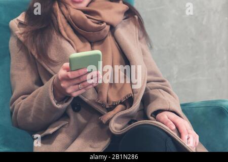 Enveloppez la femme dans un manteau marron chaud et un foulard en interaction avec le smartphone tout en étant assise dans un fauteuil doux dans un couloir de bâtiment moderne de taille haute Banque D'Images