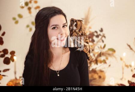Jeune femme souriante et positive vêtue de sorcière noire avec chouette sur son chagère contre un mur lumineux avec branches d'arbres Banque D'Images