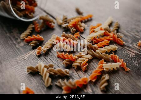 pâtes spirales brutes multicolores, orange et jaune, dispersées dans un bocal en verre sur un sol en bois sombre Banque D'Images