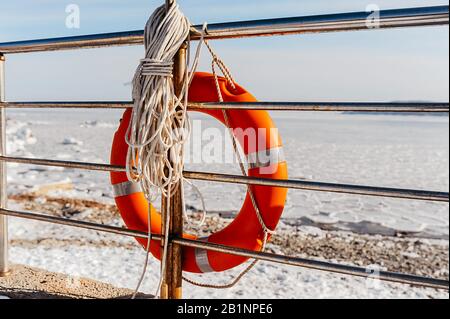 bouée de sauvetage orange vif pour aider un homme noyant, attaché avec des cordes à une rampe métallique, sur fond d'une mer gelée dans la glace Banque D'Images