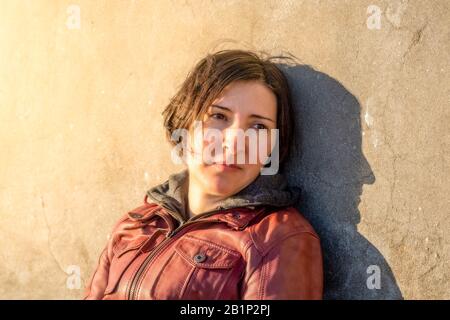 Femme brunette avec veste en cuir rouge penchée sur un mur en béton gris assis seul sous la lumière du soleil. Notion de tristesse et de solitude. Banque D'Images