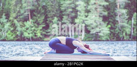 Jeune fille attrayante pratiquant le yoga, assis dans l'exercice des enfants, Balasana pose sur le lac. Le concept d'apaisement, de mode de vie sain. Panorama Banque D'Images