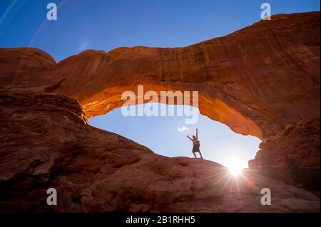 Un randonneur levant ses bras au ciel, debout sous un spectaculaire arc naturel de roche devant le soleil doré Banque D'Images
