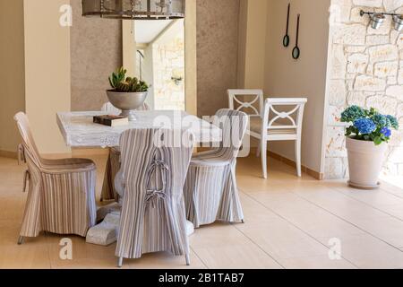 Chaises avec couvertures en tissu rayé autour de la table carrée, banc blanc, hortensia bleu dans pot à fleurs. Intérieur de salon de style méditerranéen Banque D'Images