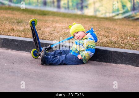 Joli petit garçon tombant de son scooter . Les enfants se blessent lorsqu'ils font du scooter. Accident de scooter en chute libre. Garçon essayant de se lever Banque D'Images