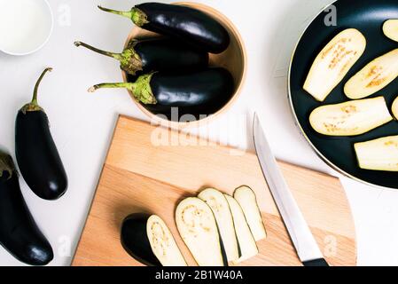 Cuisson de l'aubergine frite dans une poêle. Aubergine coupée et entière sur fond blanc. Planche et couteau en bois Banque D'Images