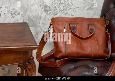 Sac d'affaires en cuir et accessoires dans la salle de travail avec mur en béton gris, table en bois et chaise en cuir. Banque D'Images