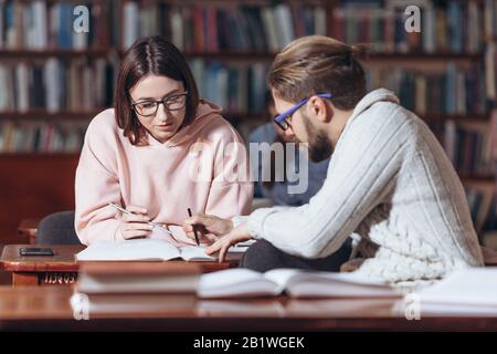 Vue latérale de l'homme barbu dans des vêtements décontractés et des lunettes expliquant les informations des jeunes étudiants à partir de livres intelligents. Homme confiant aidant jolie fille à p Banque D'Images