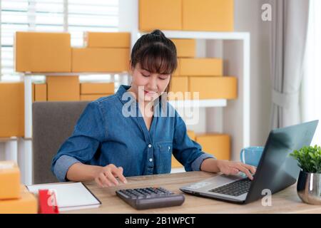 Charmante belle asiatique propriétaire adolescent femme d'affaires travailler à domicile pour le magasinage en ligne, calculer le prix des marchandises avec un ordinateur portable avec l'équipement de bureau, e Banque D'Images