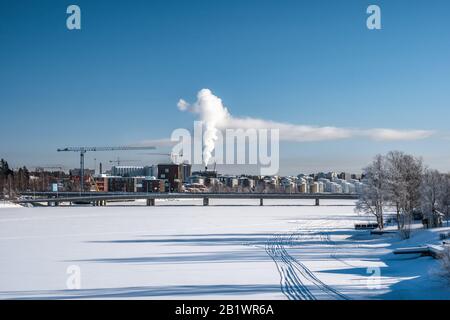 Vue de la centrale électrique de l'usine en plein centre-ville en hiver avec la vapeur qui s'élève dans un ciel bleu profond. Pont, grue, glace gelée. Journée froide. Nord de la Suède Umea Banque D'Images