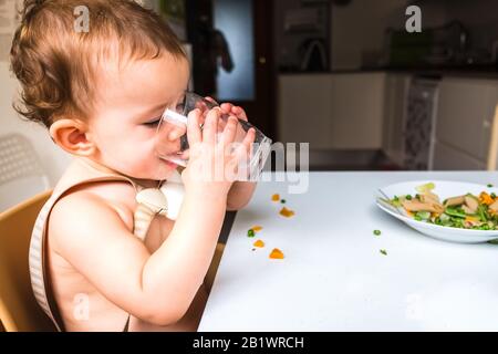 Petite fille boit de l'eau d'une tasse en verre assis sur sa chaise haute pendant le déjeuner. Banque D'Images