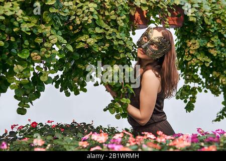 jeune femme dans une robe et un masque mystérieux parmi des fleurs et des plantes décoratives suspendues sur un fond clair Banque D'Images