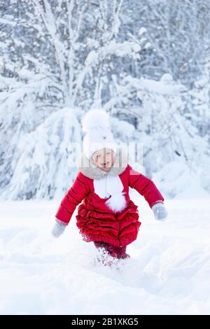 Une belle petite fille dans un manteau rouge et un chapeau blanc avec un foulard court le long d'un sentier enneigé dans un parc d'hiver. Sourire et joie des enfants sur le visage Banque D'Images