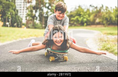Joyeux frère et soeur jouant avec le long-board en plein air - jeunes gens qui s'amusent en été dehors - jeunes, amour, famille et concept d'enfance - Banque D'Images