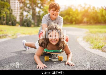 Joyeux frère et soeur jouant avec le long-board en plein air - jeunes gens qui s'amusent en été dehors - jeunes, amour, famille et concept d'enfance - Banque D'Images