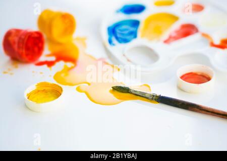 Sur la table blanche se trouvent des fournitures d'art: Une palette de plastique blanc, une brosse, des peintures gouache ouvertes, à partir desquelles la peinture s'est répandue sur la table. Banque D'Images