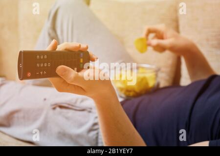 Un homme se trouve et repose sur un canapé avec une télécommande de la télévision, changer de chaîne, manger des chips de pommes de terre à partir de plats en verre. Gros plan. Banque D'Images