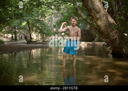 Un adolescent souriant se posant comme un bodybuilder dans les eaux peu profondes de la forêt dans le pays méditerranéen en été, soleil ambiance détendue plaisir sourire bonheur Banque D'Images