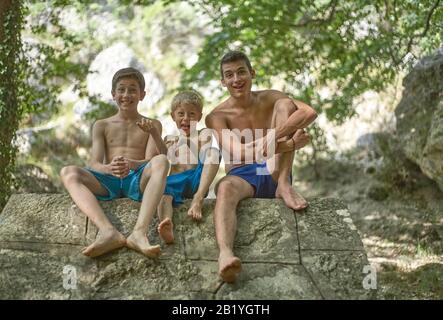 Trois frères s'amusent à faire des visages et à profiter de leur temps en été heureux souriant et détendu assis dans un short sur un rocher Banque D'Images