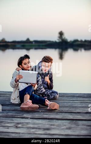 Deux jeunes petits amis mignons, un garçon et une fille qui s'amusent et font du selfie pour blogging sur une tablette numérique tout en étant assis près du lac le soir. Le Banque D'Images