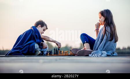 Deux jeunes petits amis mignons, un garçon et une fille qui s'amusent tout en jouant aux échecs assis avec une couverture près du lac. Les enfants jouent. Amitié. Banque D'Images