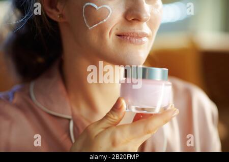 Gros plan sur une femme élégante souriante en pyjama avec coeur crème visage blanc sur joue tenant le pot cosmétique rose dans la maison moderne en hiver ensoleillé. Banque D'Images