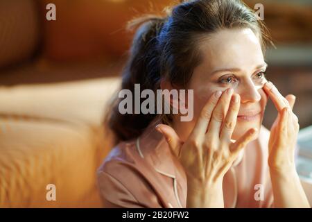 femme moderne d'âge moyen en pyjama dans la maison moderne en hiver ensoleillé appliquant la crème faciale. Banque D'Images