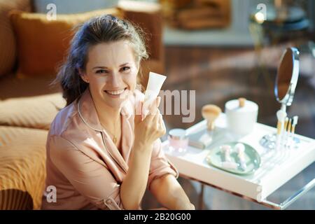 bonne femme de ménage élégante dans un pyjama montrant un tube cosmétique blanc près de la table avec des articles de toilette à la maison moderne en hiver ensoleillé. Banque D'Images