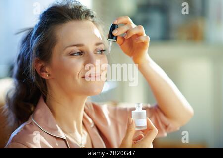 bonne femme de ménage moderne de 40 ans en pyjama dans la maison moderne en hiver ensoleillé avec élixir cosmétique. Banque D'Images