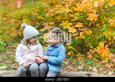 Les petites filles drôles en guerre portent assis sur le sourire de bois et se regardant l'un l'autre avec le feuillage coloré des plantes Banque D'Images