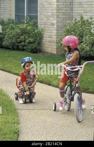 Austin, Texas - les sœurs hispaniques âgées de 5 et 2 ans sont à vélo sur le trottoir. Sœur plus âgée à vélo avec roues d'entraînement, tandis que sœur plus jeune est sur un petit tricycle. M. ©Bob Daemmrich/ Banque D'Images