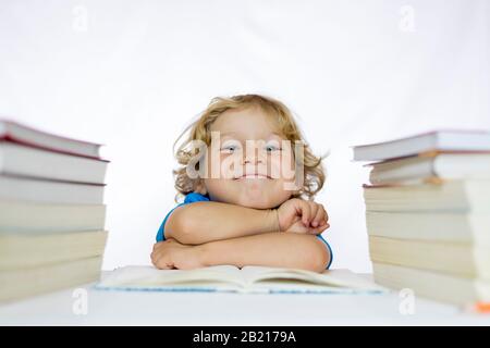 Petit enfant heureux entre 4 et 5 ans assis à un bureau étudiant avec des manuels sur sa table et avec un fond blanc Banque D'Images