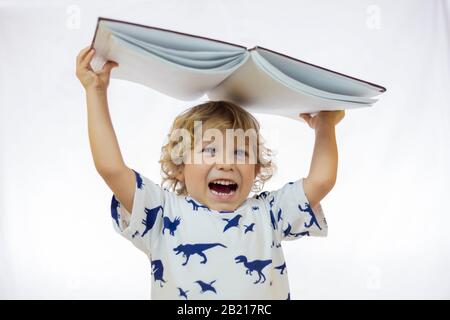 Petit garçon de 4 à 5 ans tenant un livre sur sa tête tout en souriant debout sur fond blanc Banque D'Images