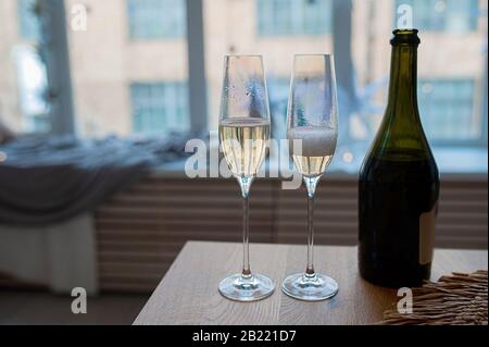 deux verres nacre avec champagne, bulles et mousse à côté d'une bouteille en verre sombre contre une fenêtre dans des tons bleus Banque D'Images